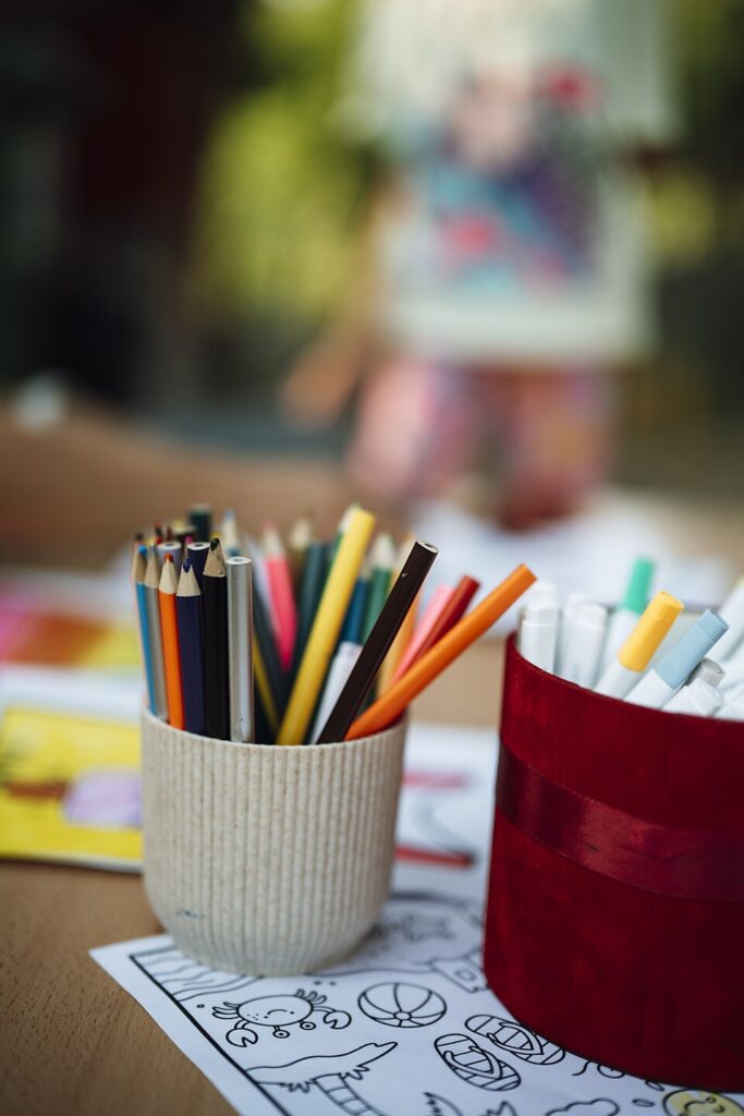 Art supplies are arranged on a table with crayons, markers, and colored pencils. A child can be seen in the background, busy with drawings during a creative activity. This file is licensed under the Creative Commons Attribution 2.0 Generic license. Source https://commons.wikimedia.org/wiki/File:Colorful_art_supplies_on_a_table_with_drawings_and_a_child_in_the_background.jpg